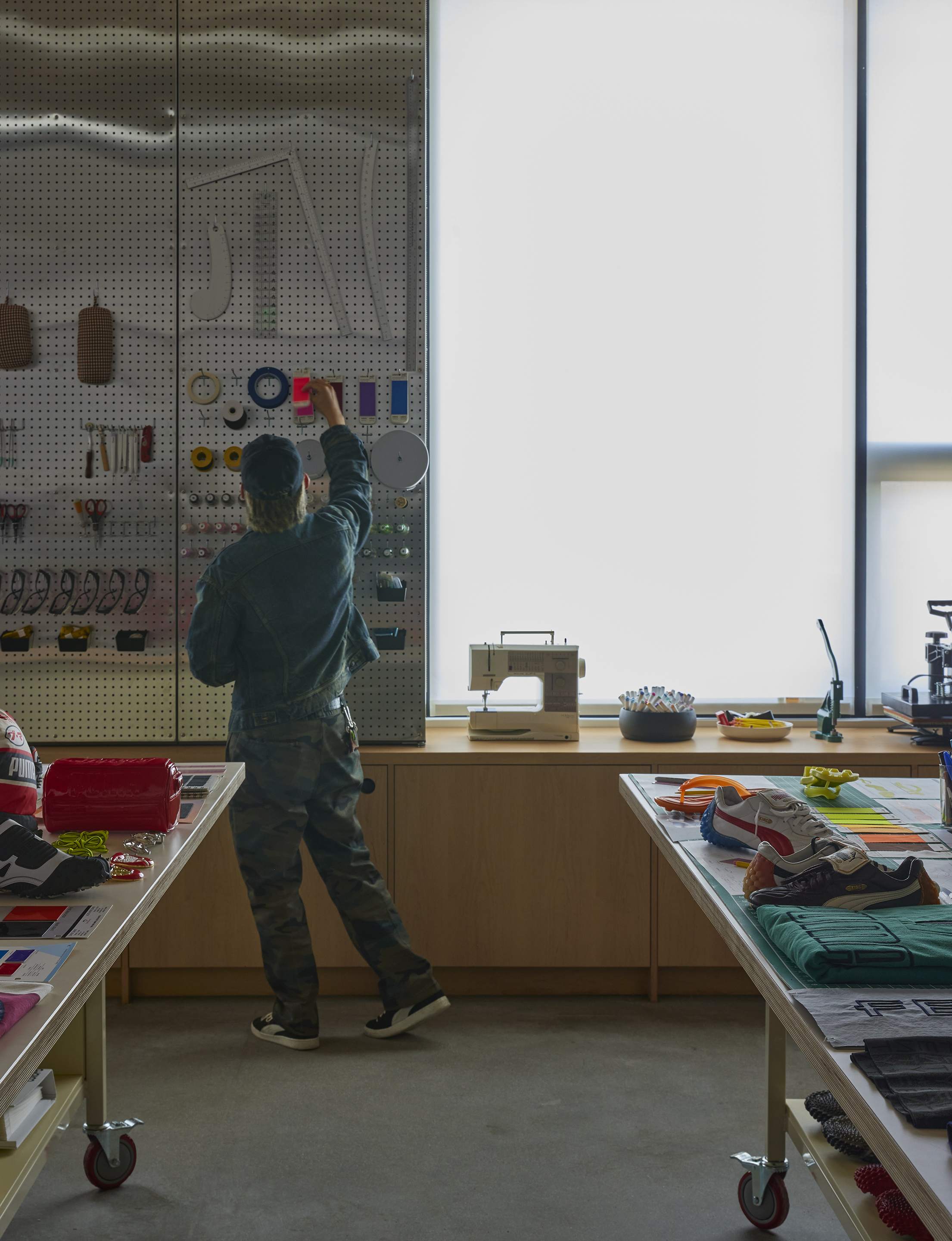 Designer at a perforated tool wall beside a sewing bench and backlit window