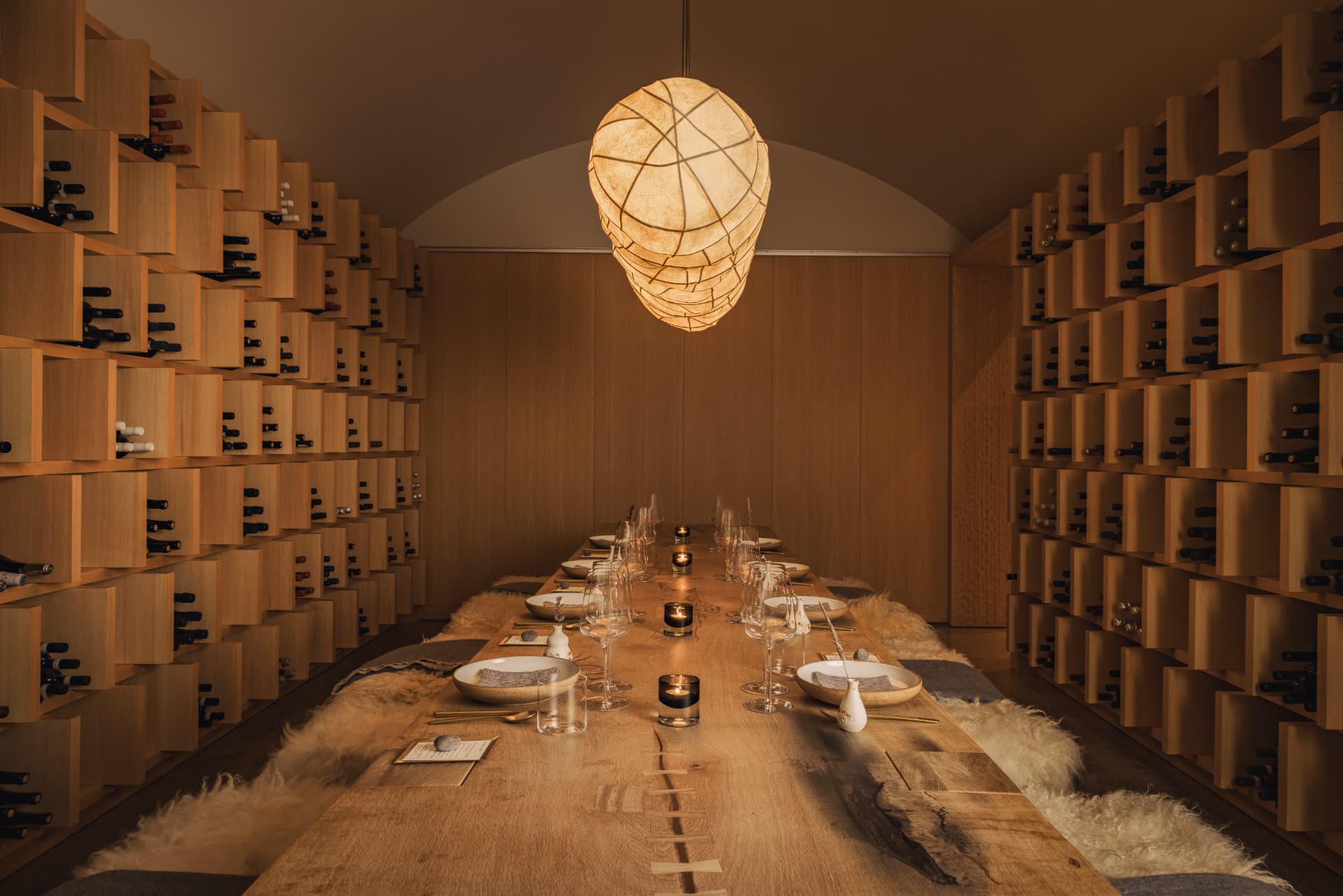 Long communal table under a woven pendant, wine wall on both sides