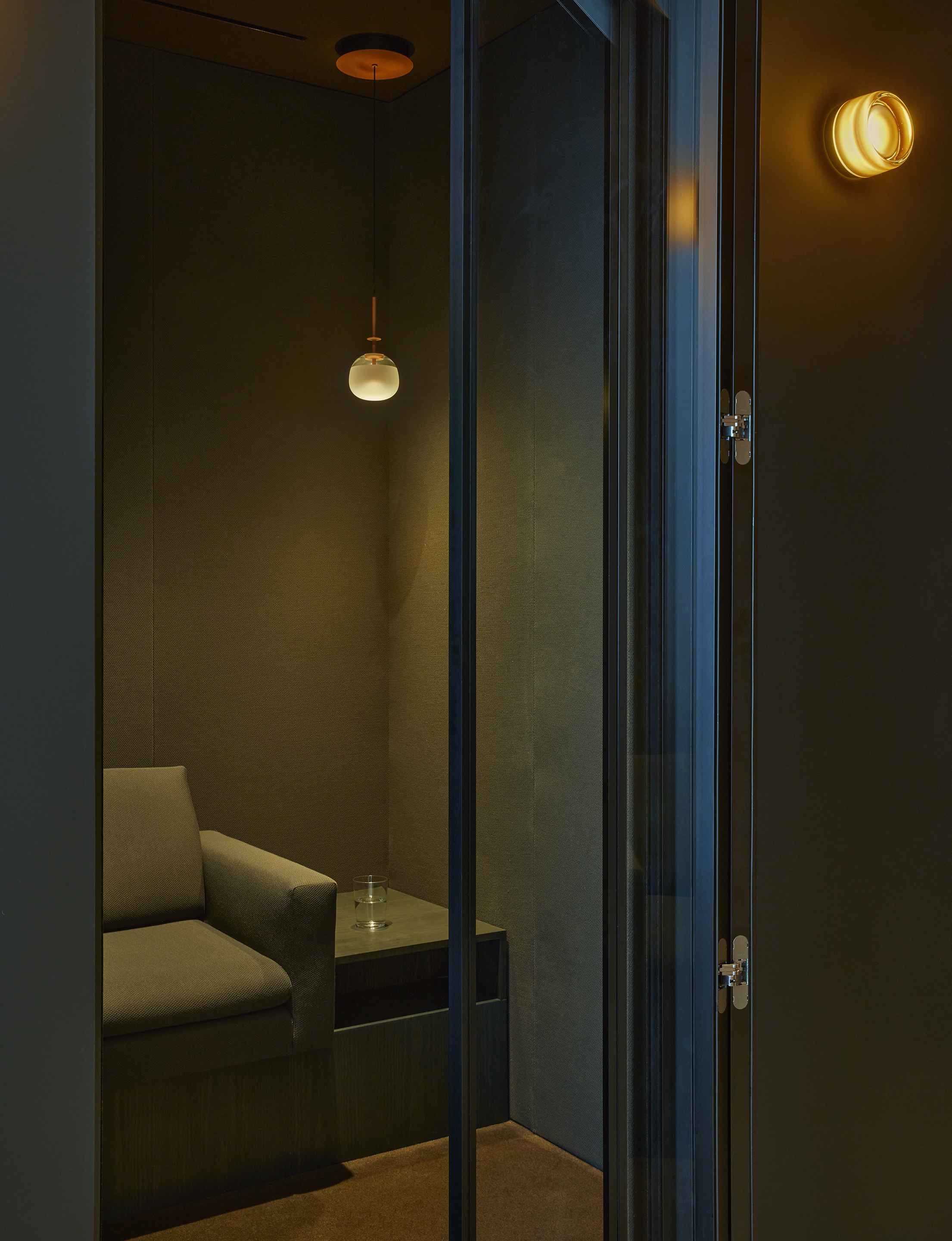 Small dark upholstered focus room seen through a glazed door under a glass pendant